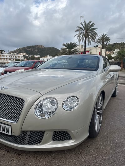 The image depicts the front-right aspect of a light champagne-colored Bentley Continental GTC convertible, parked on a dark, wet asphalt surface in Port D'andratx, Spain. The Bentley features a chrome mesh grille with the central Bentley "B" emblem, dual circular headlights with visible LED elements, and a lower front bumper incorporating mesh grilles and two visible parking sensors. A partial Spanish license plate displaying "M" followed by "MM" is affixed to the front. The vehicle's interior appears to be tan or brown, visible through the windshield, and the black convertible soft top is raised. Multi-spoke silver alloy wheels are partially visible. Water droplets are present on the car's exterior surfaces.
In the mid-ground, a red Dacia hatchback and a white vehicle are parked behind and to the left of the Bentley. Several white, multi-story buildings with Mediterranean architectural characteristics are visible behind the parked cars and scattered across the lower slopes of the distant hills. Tall palm trees are interspersed throughout the scene, particularly to the right of the Bentley and in the mid-ground. The background is dominated by a steep, forested hillside with sparse vegetation and additional white buildings. The sky is heavily overcast with significant cloud cover. No individuals are present in the visible scene.
