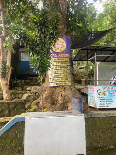 The image shows a scene in Kecamatan Manyaran, Indonesia, located at latitude: -7.826, longitude: 110.816. A large tree with a thick trunk stands in the foreground, a small stand with a sign reading "Warung Soda Asem" and "Warung Bubur" is positioned to the right of the tree. The stand is in a built-up area with a concrete wall on its right, and a small building is visible behind the tree to the left. The stand has a sign with a menu written in Indonesian. The sign lists a variety of food and drink options, including "Aneka Pop Mie," "Aneka Minuman," "Aneka Pop Ice," and "Aneka Kopi." A blue barrel is positioned to the right of the tree. A large sign is affixed to the tree with the words "Apta Pesona" and a list of Indonesian words, including "Keamanan," "Ketertiban," "Kebersihan," "Kesjukan," "Keindahan," "Keramahan," and "Kenangan." The sign appears to be promoting a place or service. The scene is dominated by a large tree, indicating the presence of a natural environment. The presence of a stand with a sign suggests a business or commercial activity. The scene seems to be taken on a bright day, with visible shadows cast by the objects, but the sky is not visible.