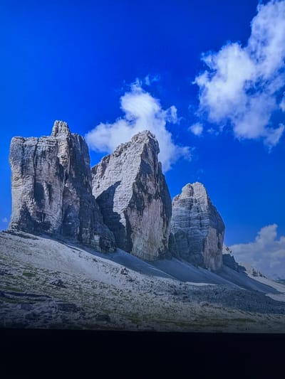 The image displays a majestic mountain range under a vibrant blue sky dotted with white clouds. The mountains are composed of layered rock formations, exhibiting textures of jagged edges and smooth surfaces. The lower slopes are covered with scree and sparse vegetation. The lighting suggests a clear day with sunlight casting shadows on the mountain faces, highlighting their rugged contours. There are no people or man-made structures visible in the image, focusing solely on the natural landscape. This scene is not located in Edmonton, Canada.