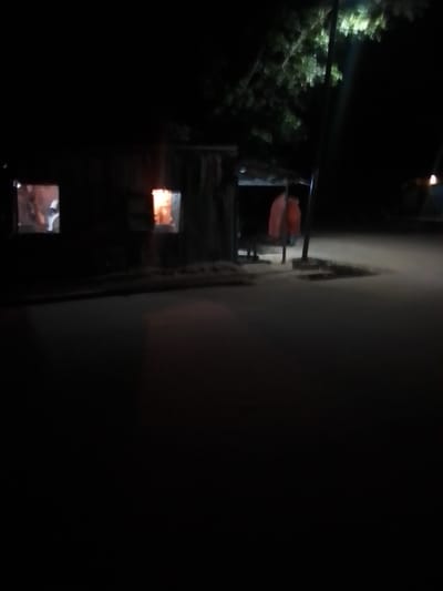 An outdoor, low-light, wide-angle shot captures a dimly lit scene at night on Liji Hill, Nigeria. In the mid-ground, a rustic, wooden structure with two visible windows is present. The left window shows a blurry interior scene with some light. The right window emanates a warm orange glow, suggesting the presence of fire or light within. Two pieces of fabric, one reddish-orange and one lighter orange, are draped over a pole to the right of the structure. A dark, smooth surface, likely a road or pathway, occupies the foreground and extends into the mid-ground, reflecting some of the ambient light. A vertical pole, possibly a utility pole, stands near the structure and the draped fabrics. Overhead, the branches of a tree are visible against the dark sky, illuminated by a bright light source. The overall atmosphere is subdued and quiet.