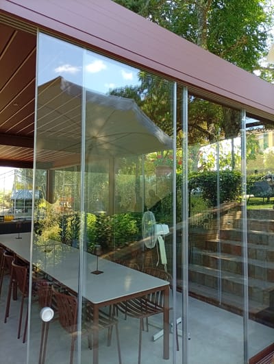 The image depicts an outdoor seating area enclosed by glass panels, likely a covered patio or veranda. A long, light-colored table is set with multiple brown chairs, suggesting a dining or gathering space. A white oscillating fan stands near the left side of the enclosure. Through the glass, a lush garden landscape is visible, complete with trees, shrubs, and a stone staircase leading upwards. The scene is bathed in natural light, indicating it is daytime. There are no people present in the image. The architectural style suggests a modern, possibly European setting, but without further context or distinctive landmarks, pinpointing a specific location within Siena, Italy, is not possible from the visual information alone.