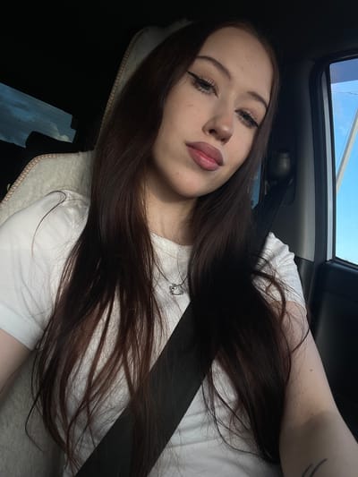 A young woman with long, dark brown hair, wearing a white t-shirt, is seen in a selfie taken inside a car. The car has a sunroof open, revealing a bright blue sky with some clouds, suggesting it is daytime with fair weather. The woman has on winged eyeliner and neutral lipstick. Her hair is spread across her shoulder and chest, and she is wearing a black seatbelt. A small, silver pendant with a 'D' on it is visible on her t-shirt. The scene appears to be a casual moment, possibly during a car ride. The interior of the car is dimly lit, contrasting with the bright sky visible through the sunroof. The image is taken from a slightly elevated angle, looking down at the woman. No text is visible in the image. The location context is Гавриловка, Russia.