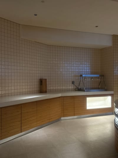 The image depicts an indoor scene, likely a food service or preparation area, with tiled walls and wooden cabinetry. A stainless steel warming unit with three heat lamps is positioned on a countertop. To the left, a small wooden dispenser is visible. The floor is tiled with large, light-colored tiles. The overall lighting suggests an artificial interior environment. The location is identified as Ras Al-Khaimah, United Arab Emirates. No people or actions are depicted in the image.