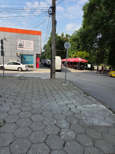 The image shows a street scene in Tutrakan, Bulgaria. A gray, hexagonal-patterned sidewalk occupies the foreground. To the left, a modern building with an orange and gray facade is visible, featuring a sign with Cyrillic text and a phone number. An air conditioning unit is mounted on the building. A white sedan is parked on the street in front of the building, next to a crosswalk. Further down the street, a white van is parked. On the right side of the street, there is a cafe with red umbrellas providing shade. Several people are seated at tables under the umbrellas, and a family with a stroller stands near the cafe. A yellow car is parked on the right side of the street. The background consists of trees and a partly cloudy blue sky. A utility pole with numerous wires is prominently featured in the center of the image.