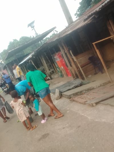 The image depicts an outdoor market or street scene in Neni, Nigeria, under an overcast sky. In the foreground, a paved surface transitions into an unpaved area bordered by a series of makeshift market stalls or small shops. These structures feature corrugated metal roofs and unpainted wooden support beams. One stall displays brightly colored clothing, specifically red, orange, and green garments, on what appear to be mannequins or forms. An empty stall contains a dark plastic chair. A utility pole with an attached solar panel and light fixture rises centrally.
Several individuals are present. In the mid-ground, a person in a green t-shirt and denim shorts stands with their back largely to the camera. Adjacent, a person in a blue top is bent over, facing two children. One child, wearing a light-colored patterned dress, holds what appear to be green fruits. A smaller child, dressed in a light blue top and shorts with pink shoes, is looking towards the camera. To the far left, partially obscured, a person in a patterned dress carries a dark bag. In the background, several adult males are visible, including one in a yellow shirt and another in a blue shirt. A motorcycle or scooter is parked on the left side of the scene. Concrete slabs, loose rocks, and a broken wooden panel are on the ground near the stalls.