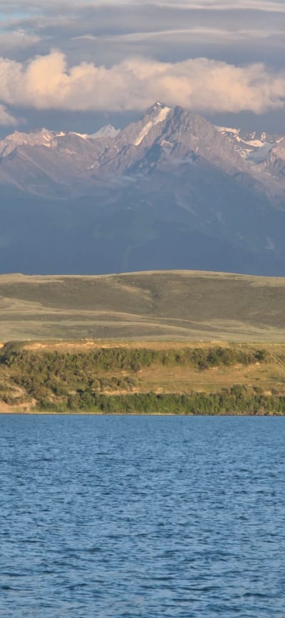The image depicts a panoramic view of a mountainous landscape with a body of water in the foreground. In the distance, snow-capped mountains rise majestically, their peaks illuminated by soft light. The middle ground features rolling hills covered in green vegetation, with a distinct ridgeline in the foreground of the hills. The foreground is dominated by a calm body of water, reflecting the sky and surrounding scenery. The sky above is partially obscured by clouds, adding a dramatic effect to the scene. The overall impression is one of serene natural beauty. This scene is located in the vicinity of the city of Pristan'-Przheval'sk, Kyrgyzstan.