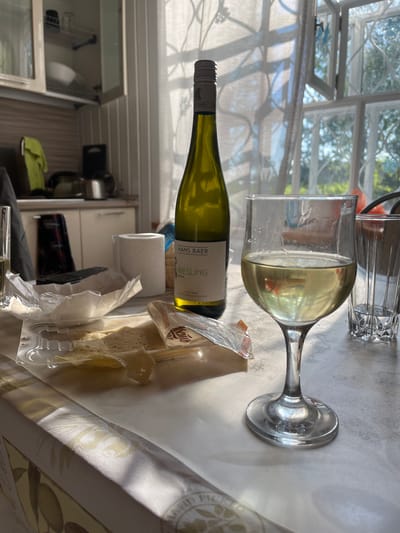 The image shows a tabletop scene in an indoor setting. A bottle of white wine, labeled "Hans Baer Riesling", lies horizontally. A wine glass containing white wine is positioned near the bottle. Next to the wine and glass, there is a package of sliced cheese on a plastic tray. A roll of toilet paper is in the background. The backdrop includes a window with sunlight filtering through curtains and a glimpse into what appears to be a kitchen with cabinets and utensils. The location in Пятница, Russia, cannot be confirmed from the image.