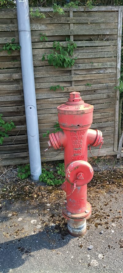 The image shows a red fire hydrant leaning against a weathered wooden fence in Mühlhausen, Germany. The fire hydrant is the primary object, displaying markings such as "DN 100 PN 16" and "DIN 3206". Its red paint is chipped in several places, revealing rust and a white undercoat. There are two caps on the top of the hydrant, each attached by a chain, and a larger outlet capped on the side. The hydrant appears to be old and possibly damaged, with one of the smaller outlets showing signs of breakage.
The fence behind the hydrant is made of vertical wooden planks, some of which are warped and weathered. Green vines and leaves are growing on and around the fence. In the foreground, a grey pipe runs horizontally across the image, partially obscuring the fence and the base of the hydrant. The ground next to the hydrant is a mixture of asphalt, dirt, and dried leaves, with some small green plants growing through it. A shadow casts diagonally across the ground from the right side of the image, suggesting it is daytime. The lighting indicates it might be late afternoon or early morning due to the long shadow. The scene appears to be outdoors in a somewhat neglected area, possibly near a street or an industrial site given the presence of the fire hydrant and the asphalt. No people are visible in the image.