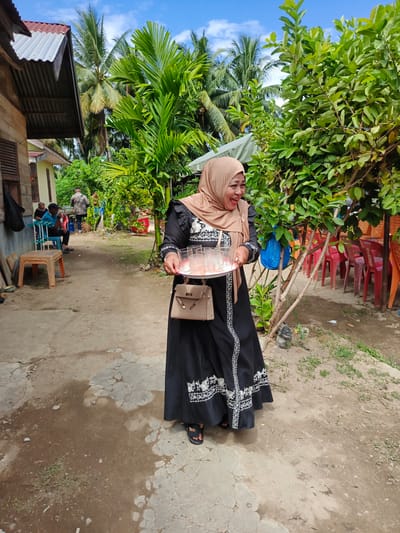 In Sangso, Indonesia, on a partly cloudy day, a woman wearing a tan hijab and a black abaya with white embroidery smiles as she carries a tray of glasses. The abaya has long sleeves and a high neckline. She also carries a textured tan handbag. The scene is outdoors, with a dirt and concrete ground. To the left, a house with a corrugated metal roof and wooden walls is visible. There are several people in the background, some seated on plastic chairs. A table is also present. A lush green tree with dense foliage is to the right of the woman, and further in the background, rows of red plastic chairs are visible. Palm trees and other greenery fill the background.