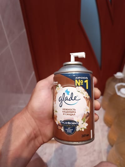 A close-up shot shows a hand holding an aerosol can of Glade air freshener. The can is predominantly brown with a dark blue band at the top, featuring the Glade logo and the text "АЭРОЗОЛЬ №1". Below the logo, in Russian, it reads "НЕЖНОСТЬ КАШЕМИРА И САНДАЛ," which translates to "Tenderness of Cashmere and Sandal." Further down, the can highlights "TRUESCENT TECHNOLOGY" and "Для ДЛИТЕЛЬНОГО АРОМАТА ЧИСТОТЫ" (For Long-lasting Aroma of Purity). The can also indicates a volume of "269 мл." The background is blurred but suggests an indoor setting with a tiled wall on the left and what appears to be large plastic containers in the background on the right. The lighting suggests it is daytime. The overall scene appears to be in Vuhlehirsk, Ukraine.