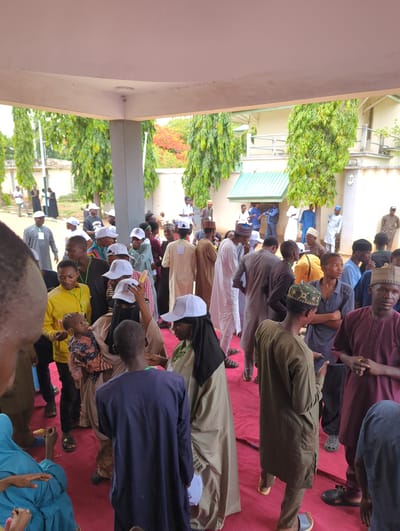 A large group of people are gathered outside under a covered area. They appear to be attending an event or ceremony. The location is Ungwan Liman, Nigeria. The people are dressed in a variety of clothing, including traditional attire and casual wear. Many people are wearing hats, some with logos. Some are seated on a red carpet or mat, and others are standing. The area is partially shaded by a building. There are trees and plants visible in the background, indicating an outdoor setting.