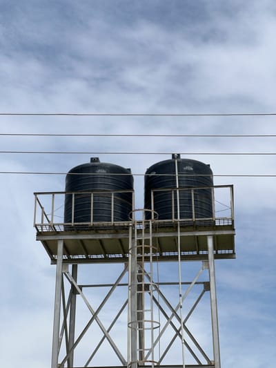 The image shows a low-angle view of a metal water tower supporting two large black water tanks. The tower is constructed of crisscrossing metal beams, with platforms and a ladder leading up to the tanks. The tanks themselves are cylindrical with ribbed sides and appear to be made of plastic. They are positioned one above the other on the tower.
The scene is set against a cloudy sky, with the clouds appearing wispy and diffused, suggesting it might be overcast or a moderately cloudy day. Several parallel wires are visible in the foreground, running vertically through the frame, indicating the presence of power lines or telecommunication cables. The image has been taken from a perspective looking upwards towards the sky.
There is some visible text on the side of the upper water tank. While partially obscured and not fully legible, it appears to be a manufacturer's label or warning.
The context provided places the location in Yali, Nigeria. The overall impression is one of infrastructure, likely serving a community in a rural or semi-urban area. The weather appears to be typical for many regions, with clouds present. No people are visible in the image. The scene is static, with the water tanks and tower simply existing in their environment.