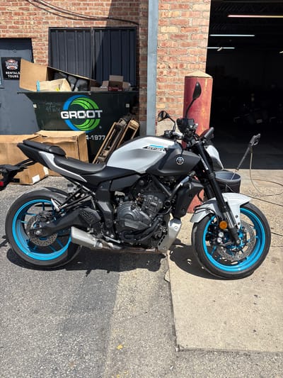 A white and black Yamaha MT-07 motorcycle is parked on an asphalt surface next to a concrete sidewalk, in front of a brick building in Des Plaines, United States of America. The motorcycle is positioned facing left, with its front wheel slightly turned. The motorcycle has bright blue rims. Behind the motorcycle, a large green dumpster is visible, with a "GROOT" logo on its side. Cardboard boxes are stacked in and around the dumpster. A red pillar stands to the right of the motorcycle. In the background, a dark garage or workshop entrance is visible, with motorcycles parked inside. The scene is brightly lit by sunlight, casting shadows on the ground.