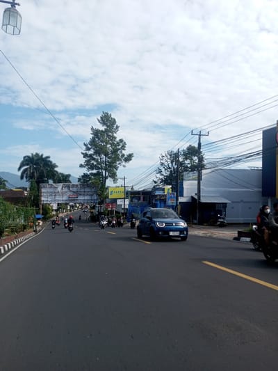 The image depicts a street scene in an urban area, likely in Southeast Asia, based on the vehicles and architecture. A blue Suzuki Ignis car is prominently positioned in the foreground, moving along a paved road with a solid yellow line marking the lane. Numerous motorcycles are also present on the road, some moving in the same direction as the car, while others approach from the opposite direction.
The background includes a mix of commercial buildings and signage. A large billboard or overpass-like structure spans the road in the distance. Utility poles and power lines are visible overhead, creating a dense network of wires. A mix of deciduous and palm trees are present on the sides of the road. The sky is partially cloudy with patches of blue. The image is taken during daylight hours. There is no indication of this location being in Zeyathiri District, Myanmar.