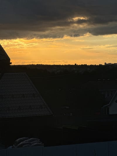 The image depicts a twilight scene with a prominent sunset sky dominating the upper portion. Dark, heavy clouds are positioned above thinner, brightly lit clouds that are illuminated by the setting sun, creating a gradient of orange and yellow hues. Below the sky, a silhouetted treeline stretches across the horizon, interspersed with a few distant structures, possibly buildings or towers, and a faint outline of city buildings on the far right. In the foreground, the dark silhouettes of rooftops are visible, one with a distinctly tiled pattern on the left, and another, lighter structure on the right. A low wall or fence is also present in the very foreground, with a pile of what appear to be sandbags or similar sacks stacked behind it. The overall impression is a semi-urban or suburban landscape at dusk, with the natural light of the sunset contrasting with the shadowed man-made structures. The location is identified as Pyachanki, Russia.