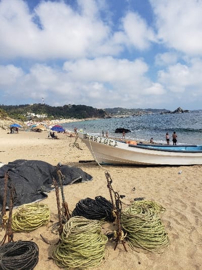 A beach scene with a white boat resting on the sand near the shore. Several bundles of rope, both light and dark colored, are scattered beside the boat. People are visible in the water and on the beach in the distance. The sky is filled with fluffy white clouds. A coastal hillside with trees can be seen along the beach.
