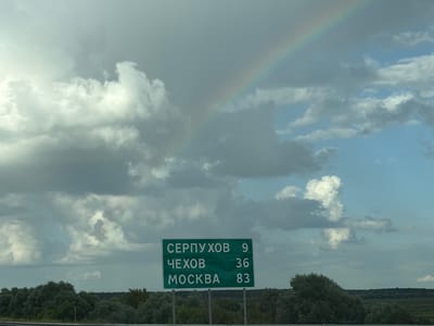 A green road sign with white Cyrillic text and numbers is positioned in the foreground, indicating directions and distances to three locations. The text on the sign reads "СЕРПУХОВ 9", "ЧЕХОВ 36", and "МОСКВА 83". Behind the sign, a lush green treeline forms the horizon, with a flat, cultivated landscape visible beyond. The sky is filled with a mix of dramatic, heavy grey clouds and patches of bright blue sky. A visible rainbow arcs across the upper right portion of the sky. The overall impression is of a landscape scene, likely in a rural or semi-rural setting, under a sky that suggests recent or impending rain. The sign itself indicates the approximate location is near Lipitsy, Russia, given the destinations listed.