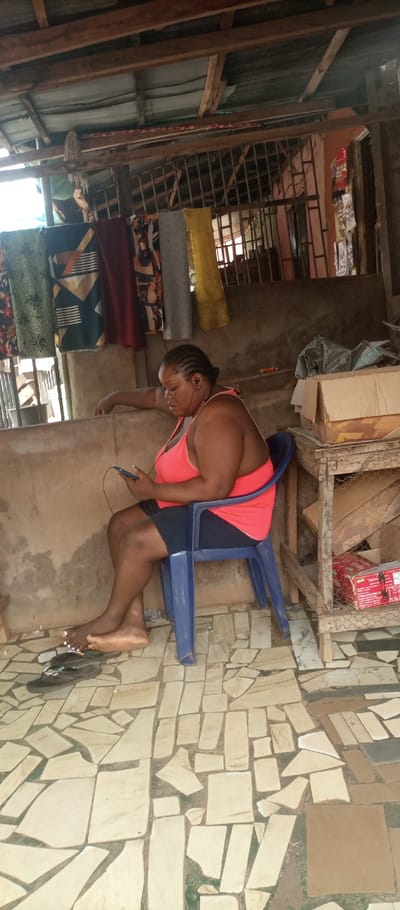 The image captures an adult female situated within an open-fronted structure in Umuoji, Nigeria. The individual, appearing to be middle-aged, has dark skin and braided hair styled back. She is dressed in a coral-colored tank top and dark shorts, and is barefoot, exhibiting white polish on her toenails. She occupies a blue plastic chair and is actively engaged with a dark-colored smartphone held in both hands, while wearing white wired earbuds. Her right forearm rests upon a low, unpainted concrete barrier that spans the foreground. A pair of dark flip-flops are positioned on the floor near her left foot.
The structure features a roof composed of corrugated metal sheets, supported by rough-hewn wooden beams and posts. Various patterned and solid-colored fabrics are suspended from lines in the mid-ground, suggesting either merchandise or laundry. To the right, a simple wooden shelving unit, partially enclosed with wire mesh, contains cardboard boxes and a red carton. The background reveals unpainted concrete walls and windows or openings secured with metal bars. The floor surface is constructed from an irregular mosaic of light-colored, fractured tile pieces set into a darker substrate.
