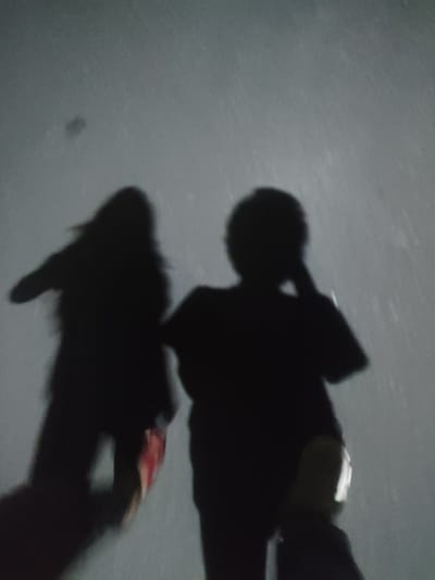 The image depicts a dimly lit scene with two silhouetted figures. The figure on the left has long, dark hair and appears to be wearing a dark top. The figure on the right is also silhouetted and appears to be holding a hand to their head. The background is a gradient of light gray to dark gray, with visible speckling that suggests rain or dust. The overall impression is of an outdoor environment, possibly at night or in low light conditions. There is no specific information in the image that allows for the identification of the location as Roranwala, Pakistan.