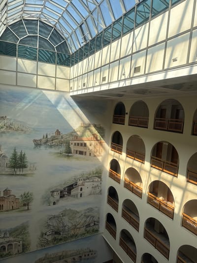 This image displays the multi-story atrium of the Hotel Arka in Skopje, North Macedonia. A prominent, vaulted glass and metal-framed skylight forms the roof, allowing direct sunlight to illuminate the interior and cast strong diagonal shadows across the walls and floor. The left wall is covered by an expansive mural depicting various historical and scenic Macedonian landmarks, including churches, monasteries, a fortified structure on a hill, traditional houses, and a stone bridge over water, rendered in an illustrative style. The right wall features a repetitive pattern of several vertically stacked floors, each characterized by a series of evenly spaced arched openings. These arched openings are fitted with light-colored wooden railings, indicating access to rooms or corridors. The dominant color scheme consists of light cream-colored walls, the varied hues of the mural, and natural wood accents. No individuals are present in the scene.