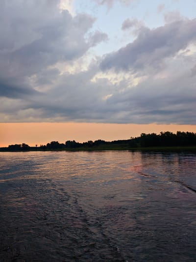 This is a vertical, medium-shot photograph of the Angara River in Kazachinskoe, Russia, during sunset. The sky is filled with dramatic, bruised-purple and grey clouds, broken by streaks of pale blue and peachy-orange light near the horizon. The light from the setting sun casts a warm glow on the lower portion of the sky and reflects on the water's surface. The river itself is dark, with ripples and small waves creating a textured, reflective surface. The far bank of the river is lined with a dense, dark silhouette of trees. The overall mood is calm and serene, with a hint of dramatic intensity from the cloudy sky. There are no people or discernible man-made structures in the image.