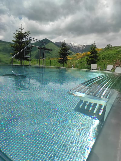 The image depicts an outdoor swimming pool with a textured bottom that reflects iridescent colors. In the background, green, rolling mountains are visible, with some peaks covered in snow. There are several evergreen trees scattered across the landscape, along with patches of yellow wildflowers. The sky is cloudy with breaks of blue. To the left of the pool, a metal structure resembling monkey bars is partially submerged in the water, with a ladder leading into the pool. In the distance, several white lounge chairs are set up on the grassy bank. The scene is set in Gudauri, Georgia, a ski resort town known for its mountainous terrain and natural beauty. The presence of the pool and lounge chairs suggests a resort or hotel setting.
