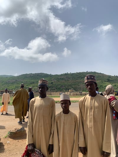 The image shows a group of men standing outdoors in Naraguta Hausawa, Nigeria. They are wearing traditional Hausa attire, including embroidered caps. A large, green hill rises in the background, with a cloudy sky overhead. Some buildings are visible near the base of the hill. The ground is dirt and gravel. The men are standing in a row, facing towards the camera.