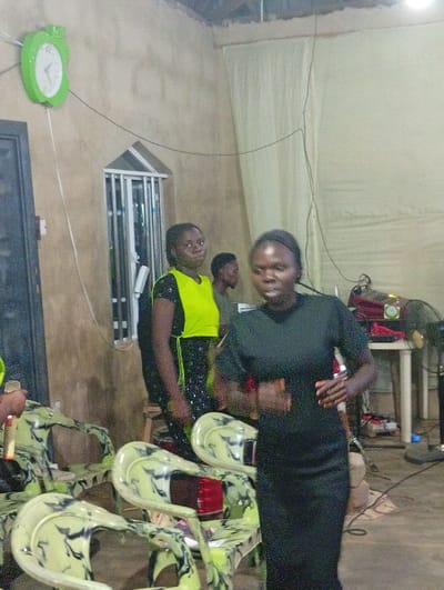 The image captures an indoor scene featuring several people and various objects, suggesting a casual gathering or event.
In the foreground, slightly to the right, a young woman with dark skin and dark hair pulled back is depicted in motion, her body angled towards the right side of the frame while her head is turned towards the viewer or slightly past. She is wearing a dark, short-sleeved dress that extends below her knees. Her hands are positioned in front of her body, and her facial expression is somewhat blurred due by her movement.
In the midground, on the left side, another young woman with dark skin stands relatively still. She is dressed in a bright, neon yellow short-sleeved top over a dark, possibly patterned, skirt or trousers. She looks off-camera to her left with a calm expression. Behind her, a young man with dark skin is partially visible, wearing a dark green or olive-colored shirt, also looking towards the left. On the far left edge of the image, part of a person's arm or hand can be seen.
The setting appears to be a room with light brown or beige walls that have a rough, unpainted texture. On the upper left wall, a prominent, bright green, apple-shaped wall clock with white hands is displayed, indicating the time is approximately 10:10. Below the clock, there is a tall, narrow, rectangular window with a distinctive peaked top and security bars or grilles. Black electrical wires are visible, running along the walls and descending from the ceiling, suggesting a functional, perhaps temporary, setup.
On the right side of the room, a light-colored table, possibly plastic, is set up with various electronic equipment, including what appears to be a projector, a speaker, and a fan. A red light is visible on one of the devices. A tall, dark-colored fan with a circular head stands on the floor near the table. A light-colored curtain or draped fabric covers part of the wall on the upper right.
Several plastic chairs with a distinctive light green and dark green/black marbled or camouflage pattern are scattered across the foreground and midground, some empty and others partially obscured. The overall lighting seems to be artificial, creating a somewhat dim ambiance. No visible text can be discerned in the image.