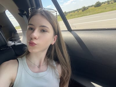 The image shows a young woman with light brown hair and fair skin, taking a selfie inside a car. She is wearing a light green ribbed tank top and a silver heart-shaped pendant necklace. Sunglasses are perched on her head. Her lips are slightly pursed, and she has subtle makeup on. She is seated in the back seat of a car, and the interior of the car is visible, including a dark-colored seat and the car door. Outside the window, a road can be seen with a green, hilly landscape under a bright, cloudy sky. The scene is likely taking place on a highway in the city of Balivka, Ukraine, given the context provided.