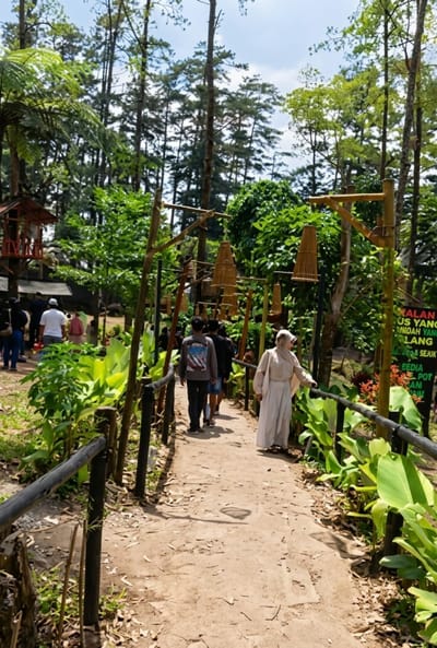 The image depicts a dirt pathway winding through a lush, green forest with tall trees overhead. Several people are walking along the path. On the left side of the path, there are large, vibrant green plants and a wooden fence. Bamboo structures with woven baskets hanging from them are visible along the path, serving as decorative elements or possibly lighting fixtures. In the background, more foliage and trees are present, suggesting a natural or park-like setting. A small wooden structure, resembling a treehouse or lookout post, is partially visible on the far left. A sign with Indonesian text is partially obscured on the right side of the path. The overall scene conveys a serene and natural atmosphere, likely in a tourist or recreational area. The location is specified as Laugumba, Indonesia.