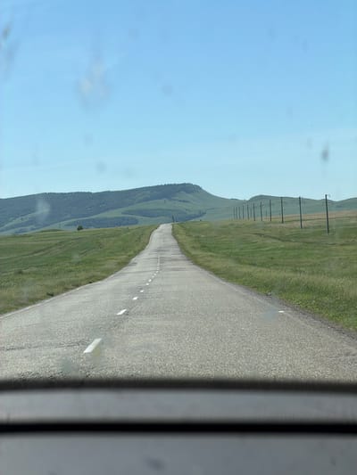 The image depicts a rural road in a hilly, grassy landscape under a clear blue sky. The road is paved and has white dashed lines marking the lanes. Power lines are visible running along the right side of the road, with poles extending upwards. In the distance, rolling hills covered in green vegetation form the horizon. A single tree stands in the middle ground to the left. The photograph appears to have been taken from inside a vehicle, looking forward through the windshield, as the bottom of the frame shows the dashboard. The location is identified as the city of Dzhirim, Russia.
