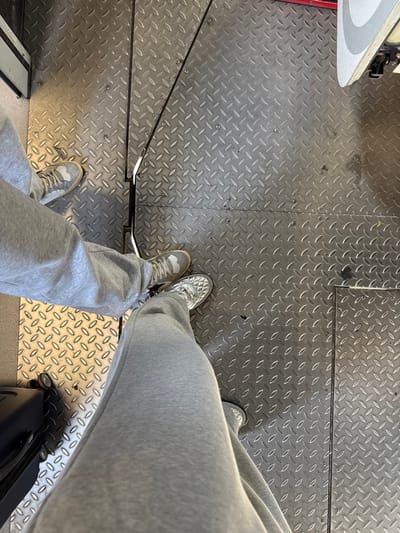 The image depicts an overhead view of two individuals standing on an aluminum diamond plate floor in a pedestrian transit area, likely a border crossing in Tijuana, Mexico. The floor consists of multiple interlocking panels secured by visible screws and features prominent black metal rails, including a central longitudinal divider.

In the foreground, the lower legs and feet of one individual are visible, clad in light grey sweatpants and silver-colored athletic shoes with a textured, possibly quilted, pattern. A portion of a black, hard-sided, wheeled suitcase is present in the bottom-left quadrant, positioned to the left of this individual's left foot.

Slightly behind and to the left, the lower legs and feet of a second individual are visible. This person is also wearing light grey sweatpants and light grey athletic shoes with white soles. Both individuals' feet are oriented generally forward. In the upper right section of the image, a white and grey structure, consistent with a vehicle or architectural fixture, is partially visible. The setting appears utilitarian and functional, common in port of entry environments.