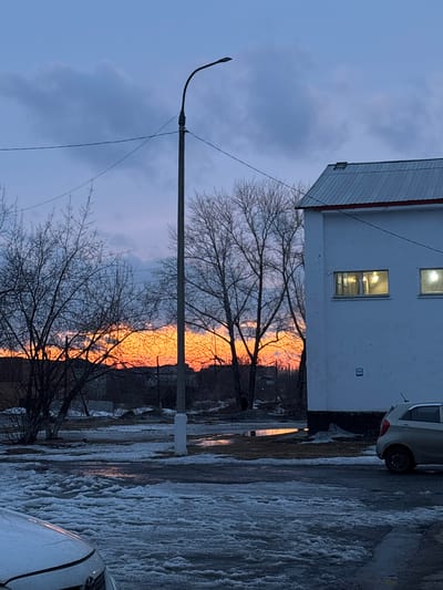 This image captures an urban scene in Tayshet, Russia, at dusk during late winter or early spring conditions. The sky exhibits a gradient from deep purple-blue overhead to a vibrant orange and red sunset glow on the horizon. The ground in the foreground and midground is covered by partially melted snow, ice, and puddles, with some puddles reflecting the sunset's colors. A white vehicle's hood is visible in the lower left. Centrally positioned is a tall, gray utility pole supporting a single streetlamp and overhead power lines. To the right, a two-story white building features a gray metal pitched roof and two horizontally rectangular windows on its upper floor, both illuminated with artificial light. A light-colored hatchback vehicle is parked facing left near the base of this building on the right side of the frame. Numerous leafless deciduous trees are interspersed across the scene, their bare branches silhouetted against the sky and the sunset. No individuals are visible.