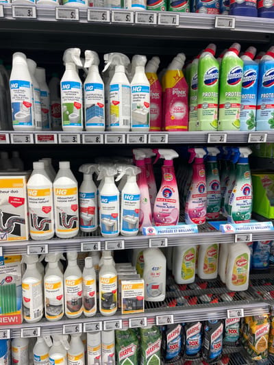 The image shows a grocery store aisle in Saint-Brice-Courcelles, France, at the coordinates 49.283, 3.999. The aisle is stocked with various cleaning products. Most of the products are in spray bottles with various colors, sizes, and labels. The aisle is organized by brands, and the top shelves are stocked with bleach-based products like Domestos. The middle shelves have various HG cleaning products for bathroom and kitchen use, while the bottom shelves are filled with Harpic toilet cleaner and other products. There is a price tag for each product, and the aisle has metal shelves with black borders. The aisle is lit by fluorescent lighting. The background is blurred out of focus.