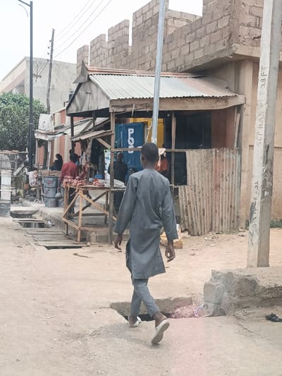 An overhead view captures a street scene in Hotoro North, Nigeria, under an overcast sky. In the foreground, a young male, dressed in a grey long-sleeved tunic and trousers with white sandals, walks away from the camera, positioned slightly left of center on an unpaved, sandy street. The ground exhibits uneven terrain, with sections of cracked concrete and dirt.
In the midground, to the left of the walking male, a rudimentary market area is visible. A wooden structure supports an open-air stall displaying raw red meat on a table. Behind this stall, a person in a reddish garment is partially visible. Further left, additional individuals are present near another stall, which features several white buckets. An open drainage channel, partially covered by wooden planks, runs parallel to the street on the left.
To the right of the meat stall, a blue shipping container, marked with "P.O." in white lettering, is incorporated into another commercial structure. This structure has a corrugated metal roof and a dark entrance opening to its right. A tall, light-colored utility pole stands in front of this section of the market.
The background on the left features a modern building with a light-colored facade and a dark roofline, along with a street light pole and overhead power lines. On the far right, a concrete utility pole displays handwritten markings including "SCP KAN LT 28FT" and "19-6-22". Adjacent to this pole is a two-story building constructed from concrete blocks, appearing unfinished or in disrepair, with exposed blockwork on the upper level and no complete roof.