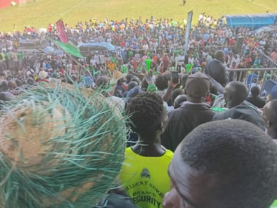 The image depicts a large, densely packed crowd of people attending an outdoor event, likely in a stadium or a similar large public space in Siaya, Kenya, during daylight hours. The perspective is from within the crowd, looking towards a central point of activity.
In the immediate foreground, a large, spherical or cylindrical object covered in coarse, green netting or strands, possibly bundled hay or natural fibers, is out of focus on the left. The mid-ground reveals numerous individuals, many seen from behind, with some holding up mobile phones, possibly recording the event. A person wearing a bright yellow t-shirt with the partial text "DR. LUCKY SUMMER BRANCH SECURITY" and an emblem is visible.
Further back, the crowd extends widely, with many people wearing green clothing, suggesting a common affiliation or theme. Green flags are also visible among the attendees. A dark-colored vehicle, possibly an SUV or pickup truck, is seen amidst the crowd, with people standing on or around it. Metal barriers or fencing separate sections of the gathering. The background shows a large grassy field, indicative of a sports ground, and parts of stadium stands.
The event appears to be a lively public gathering, possibly a political rally, a sporting event, or a festival, characterized by the large attendance and visible enthusiasm. The time of day is clear, with ample natural light suggesting a sunny day.
