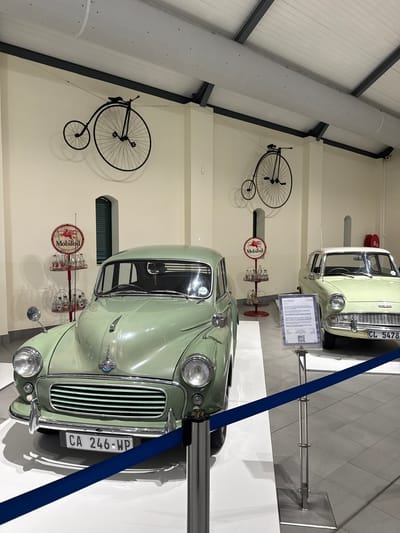 The image depicts an indoor museum exhibit featuring two vintage light green Morris Minor automobiles. One car, with license plate "CA 246-WP," is in the foreground, and another, with license plate "CL 5478," is in the background. Flanking each car are antique "MobilOil" signs mounted on stands with decorative glass bottles. Above each set of signs and bottles is a black high-wheel bicycle mounted on the wall. A blue stanchion belt partially obscures the foreground car. The scene is likely located within a museum setting in the Stellenbosch Local Municipality, South Africa, given the license plate format. The environment appears well-lit and clean, with a modern tiled floor and white walls.
