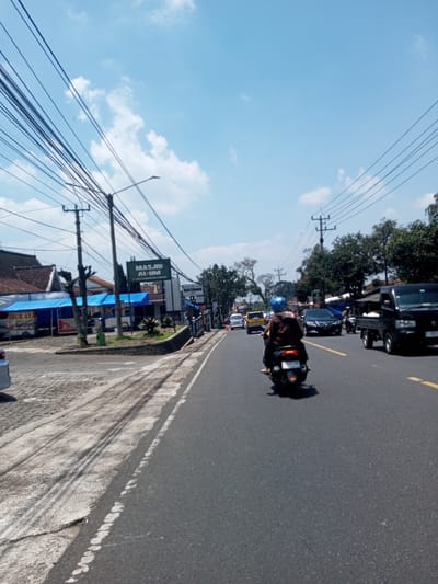 The image depicts a street scene under a bright, partly cloudy sky. An asphalt road dominates the foreground, divided by a yellow line. A motorcycle with two riders travels away from the camera. Several vehicles, including cars, a van, and a small truck, are visible ahead on the road. The left side of the road has a sidewalk and buildings with commercial signage in what appears to be Indonesian. Overhanging the street are numerous power lines connected to utility poles. A sign reads "MASJID AL-UM." The location is difficult to confirm to be in Nawng Hkar, Myanmar, as the environment, road signs, and signage are consistent with other locations in Indonesia.