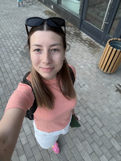 A young woman with light brown hair and blue eyes is taking a selfie outdoors. She is wearing a peach-colored ribbed t-shirt, white shorts, and bright pink sneakers. Sunglasses are perched on top of her head, and she has a black backpack on. She is holding a green object in her right hand. The background consists of a paved sidewalk made of gray rectangular bricks, a building with large windows and dark doors, and a cylindrical trash can with wooden slats. In the upper left corner, a person in blue shorts and a white t-shirt can be seen in the distance. The scene appears to be in an urban or suburban environment.