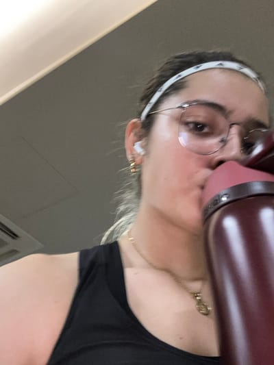 This image depicts a female subject in an indoor setting, likely in Komchén, Mexico. The subject is shown from a low angle, primarily focusing on her head and upper torso. She has light skin, dark hair pulled back, and is wearing a white athletic headband with a pattern resembling small triangles. She wears round-frame eyeglasses, a white earbud in her right ear, and a small gold hoop earring. A gold chain necklace with a small pendant is visible on her neck. She is dressed in a black tank top. The subject is actively drinking from a large, maroon-colored water bottle that obscures the lower portion of her face. The background is an indistinct light gray, likely an interior wall and ceiling, with a portion of what appears to be an air conditioning vent visible in the upper left corner.
