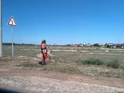 In the city of Ilyinka, Russia, a person wearing a camouflage hat and an orange and blue uniform, possibly a maintenance worker, stands in a sparsely vegetated, open field. They are holding a string trimmer and appear to be either using it or preparing to use it. A triangular road sign with a black curved arrow on a white background, indicating a sharp turn to the left, is visible on a pole to the left of the worker. In the background, a row of residential houses with multi-colored roofs stretches across the horizon under a clear blue sky. The ground in the foreground is a mix of dirt and dry grass, with some patches of green vegetation.