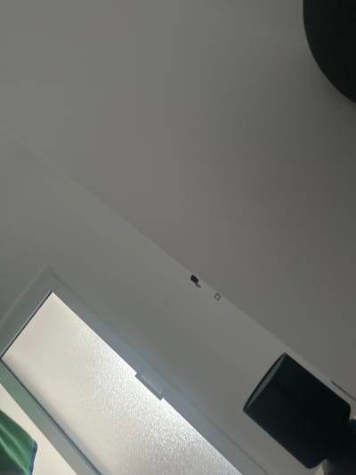 The image shows an interior view, likely a room, with a window on the left side. The window has frosted glass. To the right of the window, a black lampshade is visible, and further to the right, a dark, circular object, possibly a ceiling fixture, is partially in the frame at the very top. The walls and ceiling are painted in a light, neutral color. There are two small, dark square objects mounted on the wall above and to the right of the window. The scene appears to be taken from a low angle, looking upwards. The location, Stațiunea Zoologică Marină Agigea, Romania, does not provide specific context for the objects or room depicted.