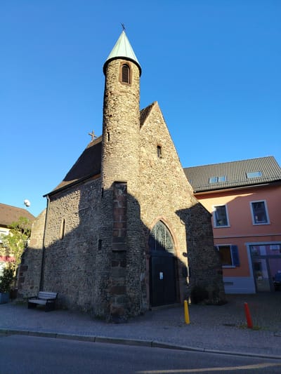 This media file shows a stone church with a tall, conical steeple topped with a green roof and a cross. The church is built with rough-hewn stones of varying sizes and colors, with some larger, darker stones used for structural elements like the corners. A small, arched window with decorative tracery is visible above the entrance, which is a large, dark double door. Sunlight casts a strong shadow of the church onto its stone facade, indicating it is likely mid-morning or late afternoon.
To the right of the church is a modern building with a peach-colored facade, featuring several windows and a grey tiled roof. Another building, partially visible on the left, has a brown tiled roof and a satellite dish on its exterior.
In front of the church, a paved area with cobblestones leads to the entrance. A dark wooden bench with a metal frame sits to the left of the church. Two brightly colored bollards, one yellow and one red, are placed on the paving. A street with a grey curb and asphalt runs along the bottom of the frame.
The scene is bathed in clear, bright sunlight under a vibrant blue sky, suggesting fair weather. There are no people visible in the image. The overall impression is one of a historic, tranquil village or town setting. The text "Achern, Germany" provides the location context.