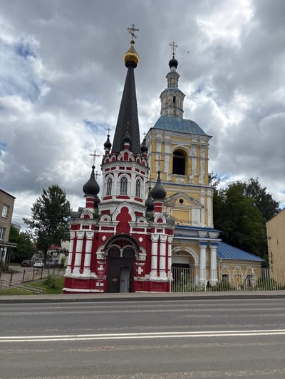 An exterior, eye-level, wide shot captures a red and white church with a tall, dark spire and a separate yellow bell tower with a blue roof, set against a cloudy sky. A paved road with white lane markings runs in the foreground, separating the viewer from a low, white metal fence enclosing the church grounds. Beyond the fence, grass and a few small white flowers are visible. To the left of the church, a brick building stands, and behind the church, trees and more buildings are partially visible. The church itself features multiple dark domes topped with crosses, and the red section has white decorative columns and arched windows. The bell tower is yellow with white trim and has arched openings. The overall scene is static and depicts a religious building within a town.