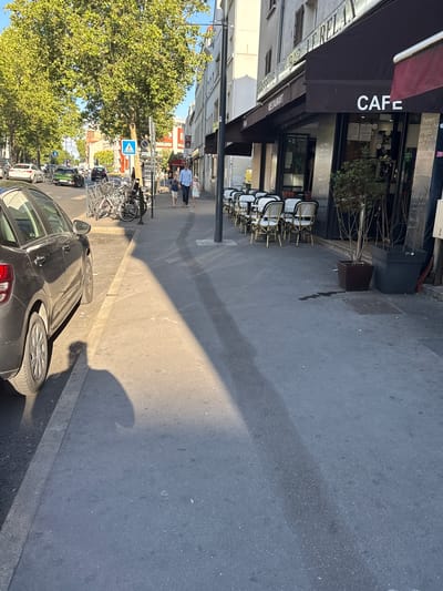 The image depicts a street scene in Colombes, France. On the right side of the image is a brasserie and bar named "LE RELAX" with outdoor seating. The brasserie has a dark awning with "BAR BRASSERIE LE RELAX" and "RESTAURANT" written on it. White chairs with woven seats and backs are set around small tables on the sidewalk in front of the establishment, some of which are occupied by potted plants. The word "CAFE" is prominently displayed in large white letters above the entrance. Further to the right, a dark rectangular object, possibly a trash receptacle or utility box, stands next to a sign with text.
On the sidewalk in front of the brasserie, two children, a boy and a girl, are walking away from the camera. They are accompanied by an adult male who is also walking away. In the background, on the left side of the street, cars are parked and driving. One green car is visible driving in the distance, and two white cars are parked on the side of the road. A pedestrian crossing sign with a blue background and a white pedestrian symbol is visible on a pole in the middle of the street.
A dark-colored car is parked on the left side of the image, partially obstructing the view of the sidewalk and street. The car's rear is visible, showing its taillight and wheel. The scene is illuminated by sunlight, casting long shadows across the sidewalk and street. The environment appears to be a typical urban street with buildings lining the right side and trees and parked cars on the left.