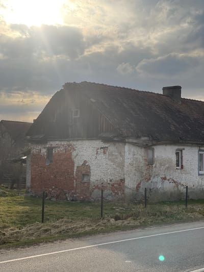 The image depicts an old, dilapidated building in a rural setting. The building has a steeply pitched, dark red tiled roof and a chimney. The exterior walls are a mix of exposed red brick and white plaster, with significant deterioration and missing sections revealing the underlying brick. There are several small windows with white frames visible on the plastered section of the wall. The building appears to be adjacent to a road with a white line marking the edge. A grassy area is present between the building and the road. The sky is cloudy, with the sun shining through, creating a hazy and bright effect. The image suggests a state of disrepair and neglect. The location is Rovnoye, Russia.