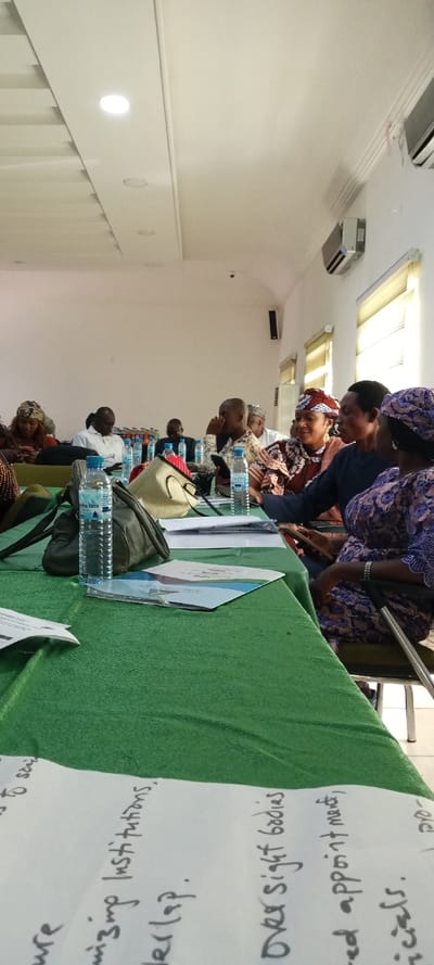 The image captures an indoor meeting or workshop setting in Yola, Nigeria. Approximately ten adults are seated around a long table draped with a green tablecloth.

In the foreground, a plastic bottle of "Elo Inna" brand water, a dark shoulder bag, and a lighter-colored woven bag rest on the green tablecloth. Several papers and documents are distributed on the table, including one in the bottom left corner with legible handwritten notes such as "organizing institutions, overlap," and "Oversight bodies, appointment."

Mid-ground, on the right side of the table, three women are engaged in interaction. One woman, wearing a red and brown patterned headwrap and dress, is looking towards a woman in a dark blue top. A third woman, wearing a purple patterned dress and bracelets, holds a mobile phone. Further along the table, other participants, including men and women, are visible, some observing the room, others focused on personal devices or documents. Multiple additional bottled water containers are present on the table.

The room features plain white walls and a white ceiling equipped with several recessed circular light fixtures. Two white, wall-mounted air conditioning units are visible on the right wall, above vertical window blinds. Small, black rectangular speakers or sensors are also affixed to the white walls.