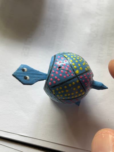 The image displays a small, handcrafted figurine, likely made of wood or a similar material, set against a plain white paper background. The central subject is a stylized blue creature, resembling a turtle or an aquatic animal, positioned vertically in the frame.

The creature's body is rounded and dome-shaped, painted a light blue. Its shell is intricately decorated with a pattern of black lines that divide it into segments. Within these segments, small, raised dots alternate between vibrant yellow and bright pink. A small, dark hole or indentation is visible on the left side of the shell, within one of the pink-dotted sections.

Extending downwards from the main body is a flat, pointed tail-like or head-like extension, also painted blue. This lower section features two small, round white eyes with black pupils, giving the creature a whimsical appearance. A pointed fin or tail protrudes upwards from the top of the main body.

The figurine is partially obscured at the top and right by the presence of human fingers, suggesting it is being held or positioned on the paper surface. The background paper appears to be standard white office paper, with very faint, indistinct textual imprints or watermarks visible upon closer inspection, but no legible words. A dark, thin object, possibly a pen or ruler, is partially visible along the right edge of the frame. The lighting indicates an indoor setting.