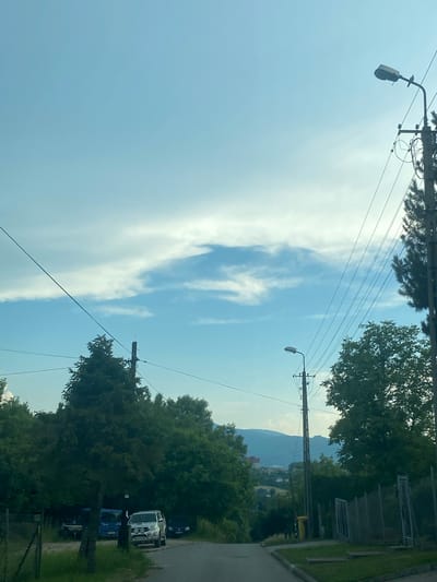 The image depicts an outdoor scene, oriented vertically with the sky dominating the upper portion. The sky is a light blue with wispy white clouds, suggesting it is daytime with fair weather.
Prominently featured are utility poles with power lines stretching across the frame. One pole in the foreground has a street lamp attached to it. Further in the background, a line of trees lines a paved area, possibly a road or a parking lot. Parked vehicles are visible amongst the trees, including a white van and a dark blue van. Beyond the trees and vehicles, a gentle slope leads up to a hazy mountain range under the bright sky.
The overall impression is of a suburban or semi-rural setting during the daytime. There are no people visible in the image. No discernible text is present. The orientation of the image is such that the top of the frame appears to be the sky, and the bottom is closer to the ground.