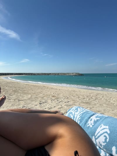 The image presents a first-person perspective shot from a sandy beach in Ghadeer Al Tayr, United Arab Emirates. In the immediate foreground, the upper thighs and lower torso of one individual are visible, exhibiting a tanned skin tone. A light blue towel with a white coral pattern is positioned under the individual's right leg. The subject is in a reclined position, facing the sea. The middle ground consists of a wide, light-colored sand beach that curves towards a prominent man-made rock barrier, or breakwater, extending into the water. Beyond this barrier, the sea displays a turquoise hue, with small waves breaking near the shore. Several small, white buoys are visible in the water in the mid-distance. The background features a clear, bright blue sky with sparse, wispy white clouds. The scene is illuminated by strong daylight, indicating clear weather. No other individuals or specific actions beyond reclining are discernible.